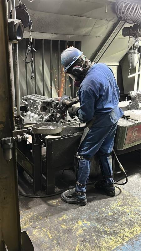 Trabalhador em esmerilhamento de bloco de motor, usando máscara, protetor auricular e avental